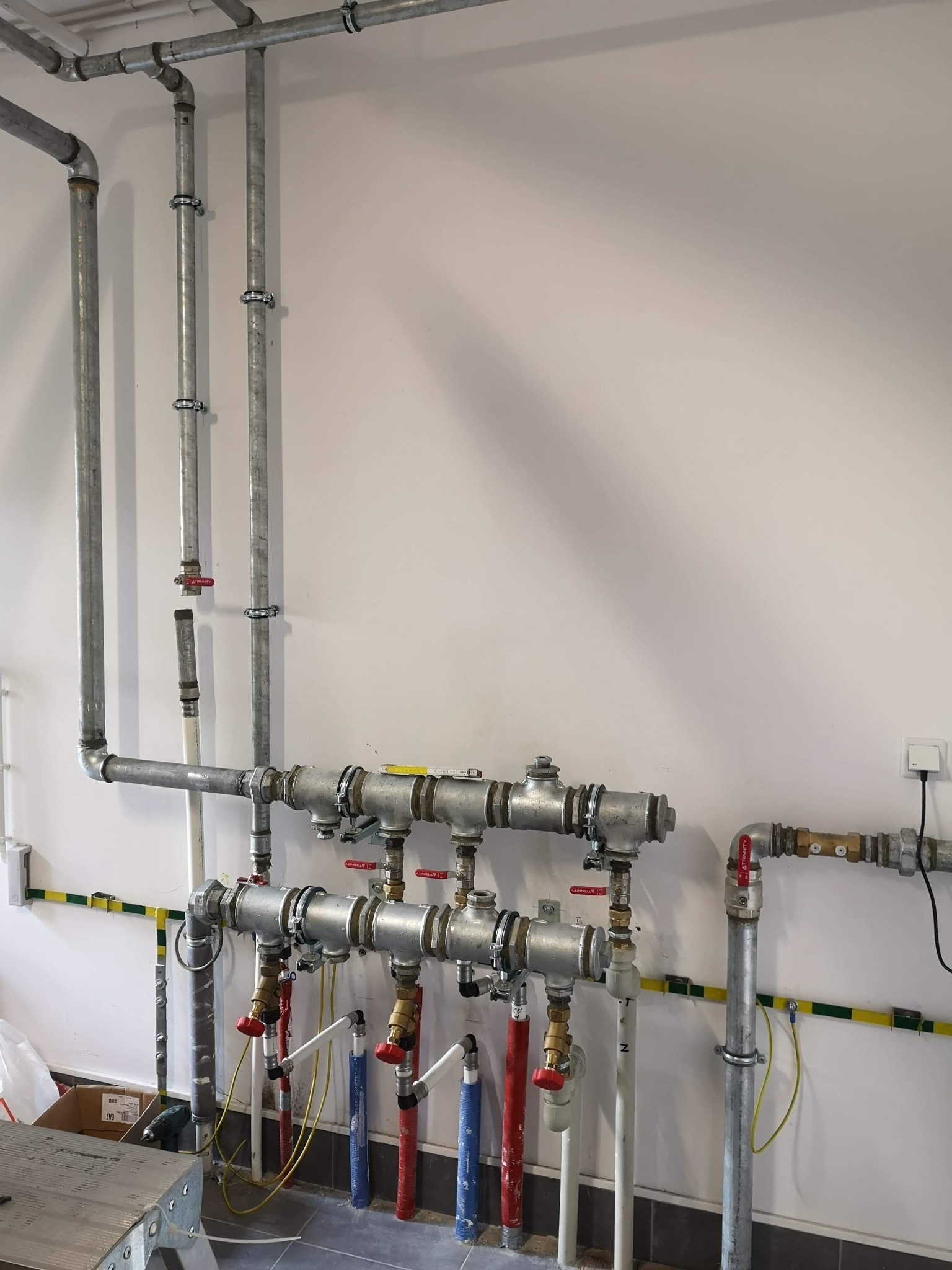 Szare metalowe rury instalacji grzewczej z zaworami z czerwonymi pokrętłami, przymocowane do białej ściany za pomocą srebrnych obejm, widoczne elementy instalacji elektrycznej i fragment drabiny.