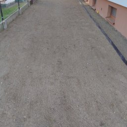 LATO-GARDEN - Przygotowany teren pod trawnik z widocznymi śladami po maszynach, obrzeżami i fragmentem budynku w tle. Grunt wyrównany, gotowy do dalszych prac.