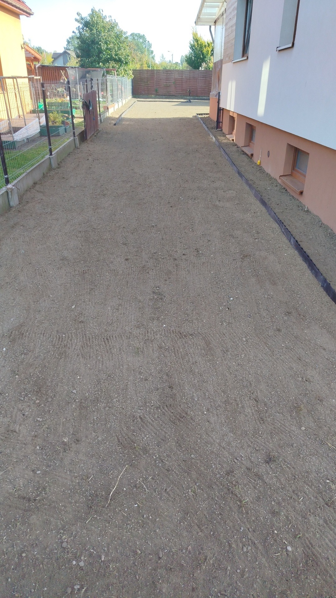 Przygotowany teren pod trawnik z widocznymi śladami po maszynach, obrzeżami i fragmentem budynku w tle. Grunt wyrównany, gotowy do dalszych prac.