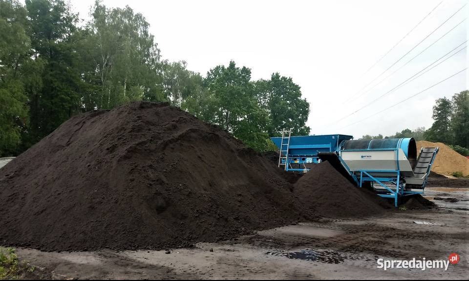 Góra ciemnej ziemi obok przesiewacza do gleby z drabiną, na tle drzew i linii energetycznych. Ślady opon na ziemi. Przesiewanie ziemi.