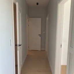 Korytarz w jasnych barwach z drewnianą podłogą, białymi drzwiami z czarnymi klamkami i szarymi ścianami, oświetlony punktowymi lampami sufitowymi.