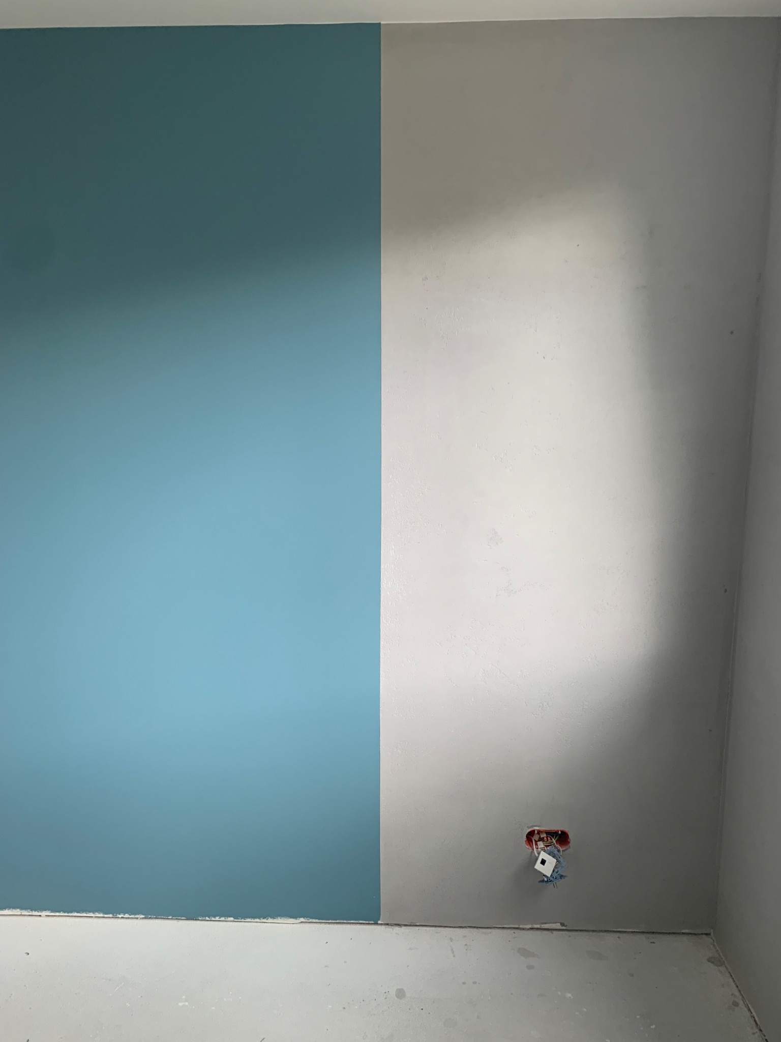Ściana w rogu pokoju, pomalowana w połowie na błękitno, w połowie na szaro, z widoczną puszką elektryczną bez osprzętu.