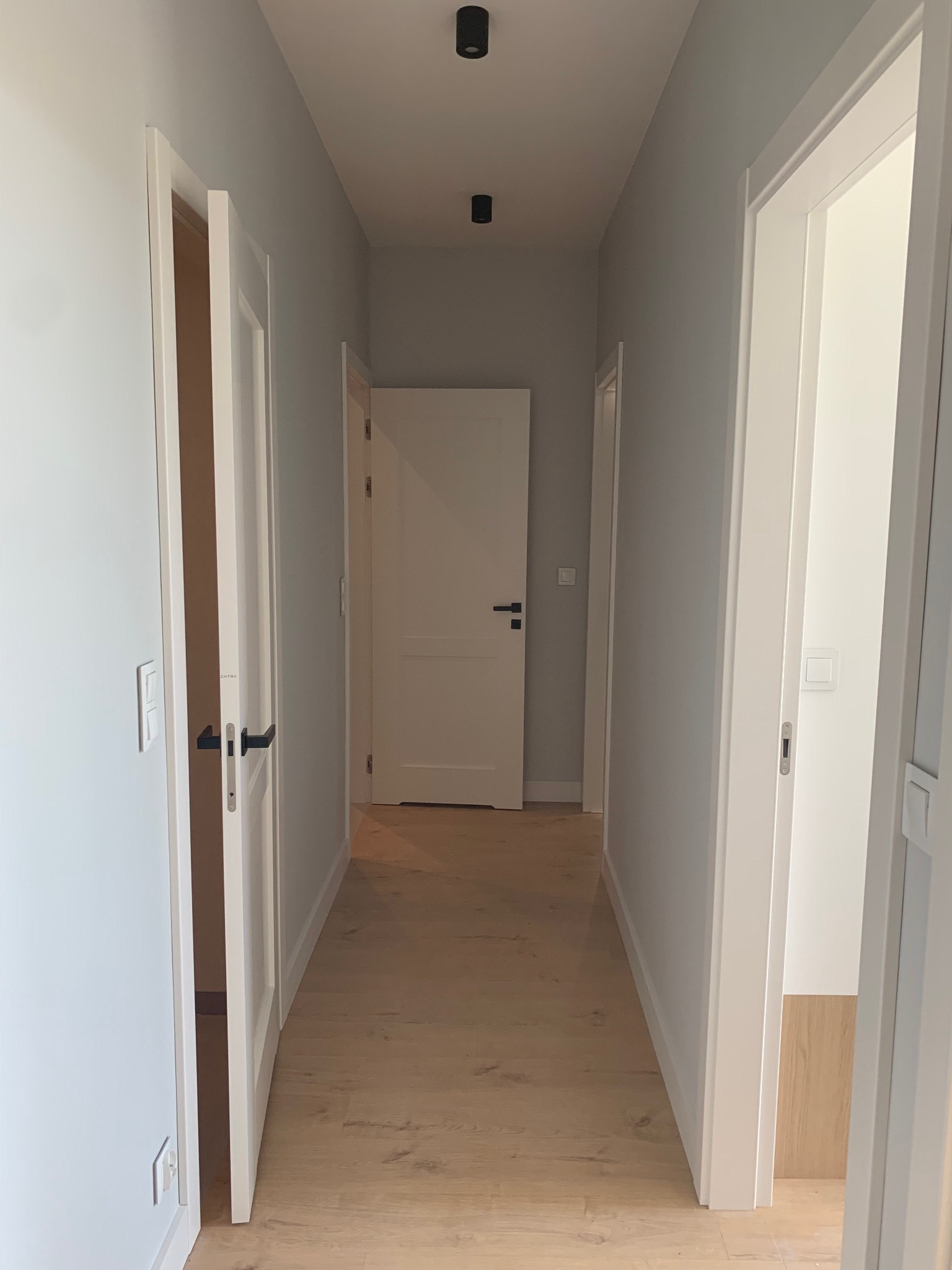 Korytarz w jasnych barwach z drewnianą podłogą, białymi drzwiami z czarnymi klamkami i szarymi ścianami, oświetlony punktowymi lampami sufitowymi.