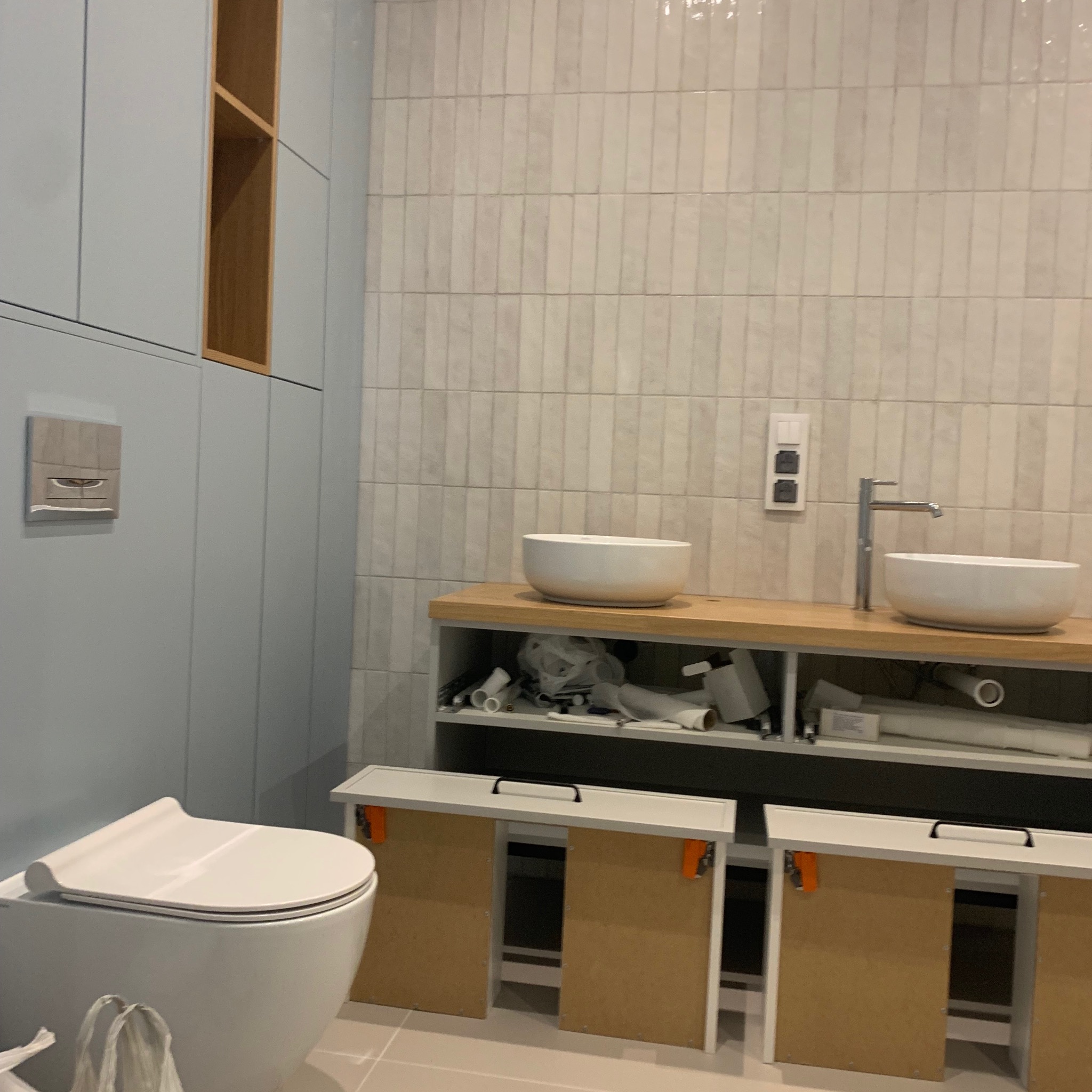 Łazienka w trakcie prac wykończeniowych: wisząca toaleta, umywalki nablatowe, armatura, szafki pod umywalkami z zamontowanymi drzwiczkami, płytki ścienne w odcieniach bieli, widoczne materiały...