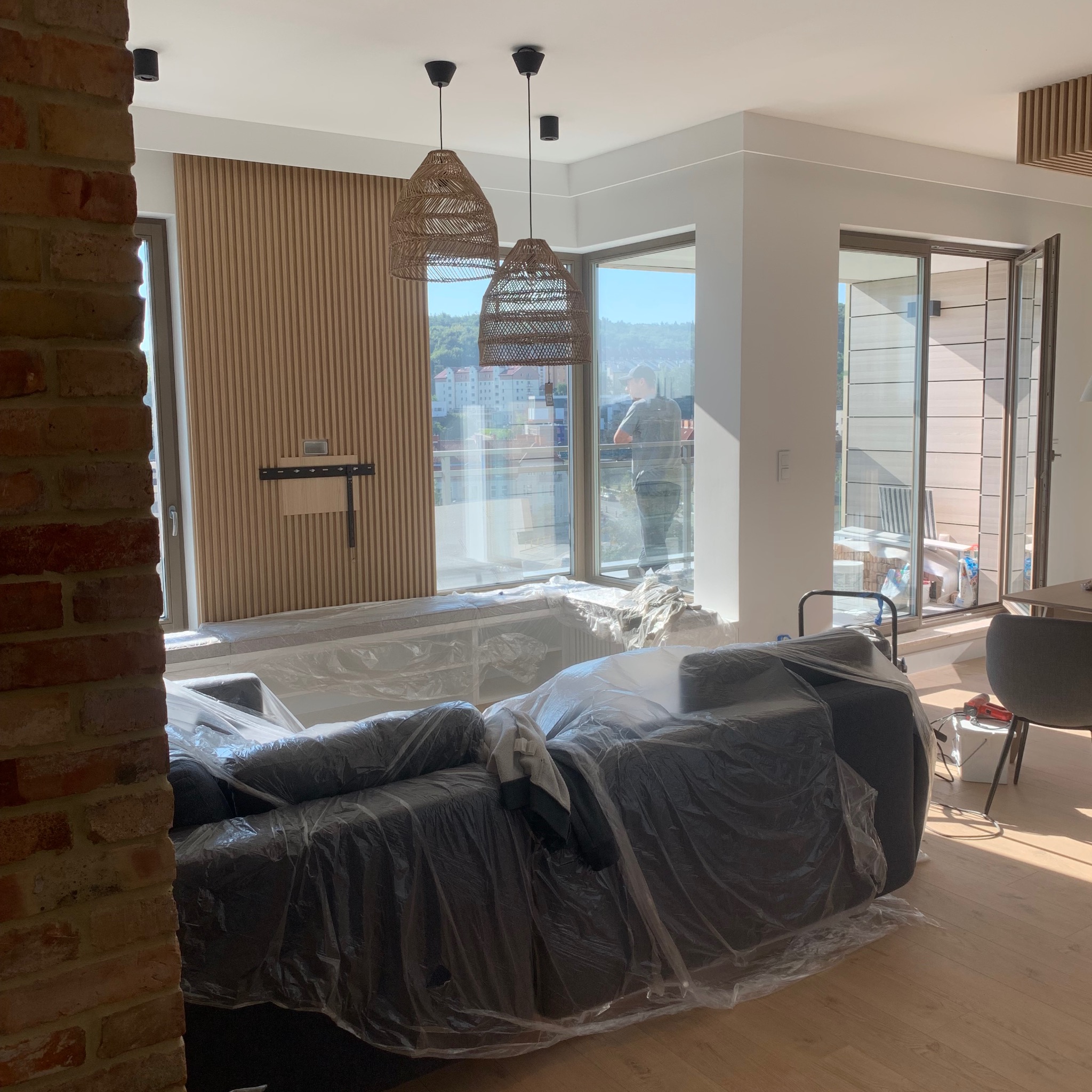 Wnętrze mieszkania w trakcie prac wykończeniowych: zabezpieczony folią mebel, drewniane panele ścienne, dwa ażurowe abażury lamp wiszących, widok na balkon z osobą w tle.