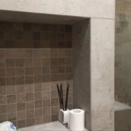 Wnęka w kabinie prysznicowej wyłożona mozaiką w odcieniach brązu, z dekoracyjnym odświeżaczem powietrza, patyczkami zapachowymi i rolką papieru toaletowego, obok szklana ściana prysznica...