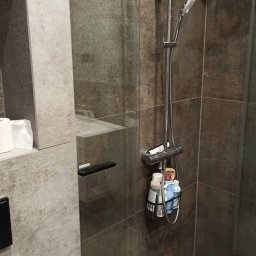 m4home Marcin Szmidt - Szklana kabina prysznicowa z chromowaną armaturą i półką na kosmetyki, wykończona płytkami imitującymi kamień w odcieniach szarości i brązu, z widocznym fragmentem toalety i wnęką z rolkami papieru...