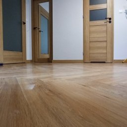 m4home Marcin Szmidt - Ułożona parkietowa podłoga w jodełkę francuską, widoczne trzy drewniane drzwi z przeszkleniami i czarnymi klamkami, białe ściany i listwy przypodłogowe.