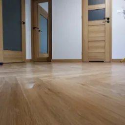 Ułożona parkietowa podłoga w jodełkę francuską, widoczne trzy drewniane drzwi z przeszkleniami i czarnymi klamkami, białe ściany i listwy przypodłogowe.