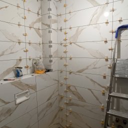 m4home Marcin Szmidt - Łazienka w trakcie układania płytek marmuropodobnych z użyciem krzyżyków dystansowych i listew narożnych. Widoczna drabina i narzędzia glazurnicze.