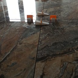 m4home Marcin Szmidt - Połyskujące, marmurowe płytki podłogowe z systemem poziomowania płytek. Widoczne pomarańczowe kliny i białe klipsy. Płytki ułożone równo, z zachowaniem spoin.