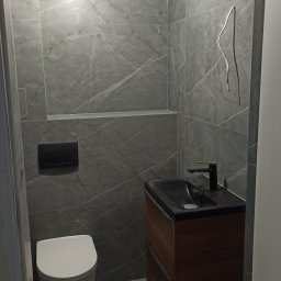 m4home Marcin Szmidt - Nowoczesna łazienka z szarymi płytkami imitującymi marmur, podwieszana toaleta i drewniana szafka z czarną umywalką. W tle wnęka w ścianie.