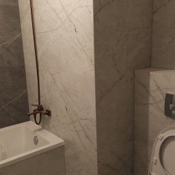 m4home Marcin Szmidt - Wykończona łazienka z marmurowymi płytkami, brązową armaturą i białą ceramiką. Widoczna wanna, toaleta i bateria prysznicowa. Całość utrzymana w stonowanej kolorystyce.