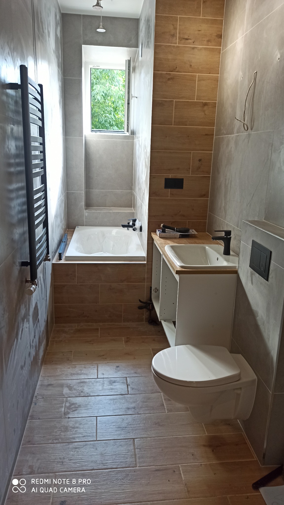 Wnętrze łazienki w trakcie wykańczania, z widoczną wanną, umywalką z szafką bez frontów, wiszącą toaletą i grzejnikiem drabinkowym, wykończone płytkami imitującymi drewno i szarym gresem.