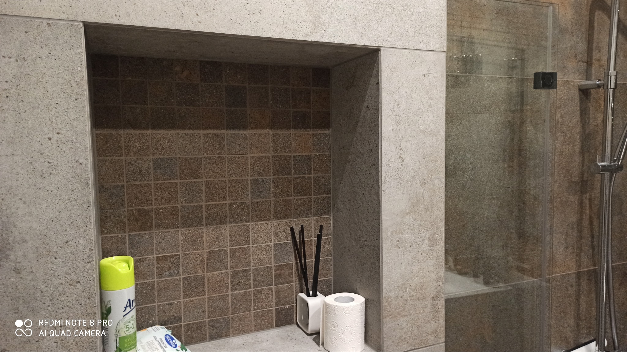 Wnęka w kabinie prysznicowej wyłożona mozaiką w odcieniach brązu, z dekoracyjnym odświeżaczem powietrza, patyczkami zapachowymi i rolką papieru toaletowego, obok szklana ściana prysznica...