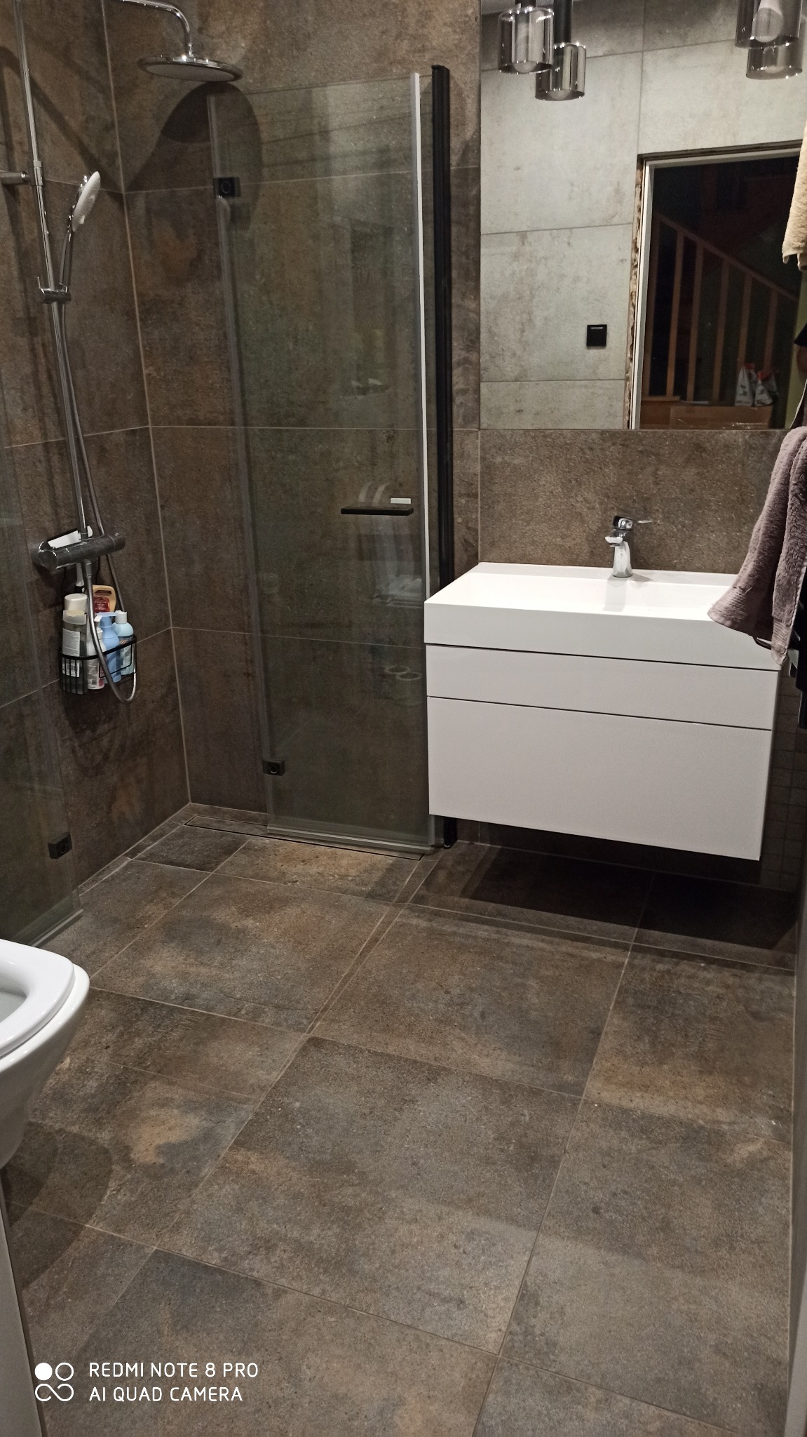 Nowoczesna łazienka z kabiną prysznicową typu walk-in, brązowymi płytkami imitującymi kamień, białą szafką pod umywalką i lustrem z widocznym odbiciem schodów.