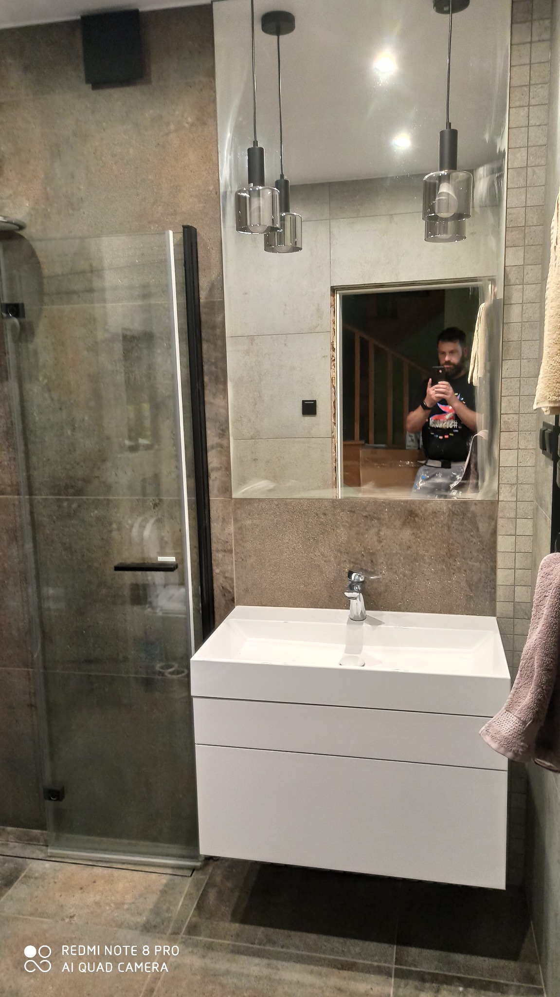 Nowoczesna łazienka z kabiną prysznicową ze szkła, białą szafką pod umywalką i lustrem, w którym odbija się osoba robiąca zdjęcie.