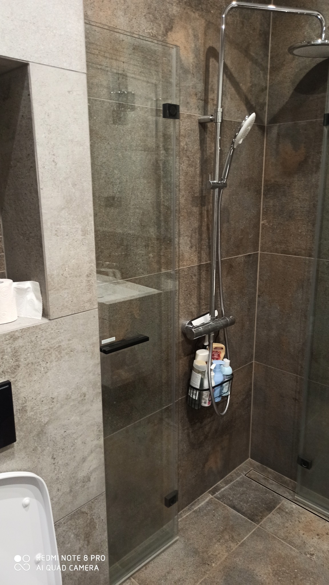 Szklana kabina prysznicowa z chromowaną armaturą i półką na kosmetyki, wykończona płytkami imitującymi kamień w odcieniach szarości i brązu, z widocznym fragmentem toalety i wnęką z rolkami papieru...