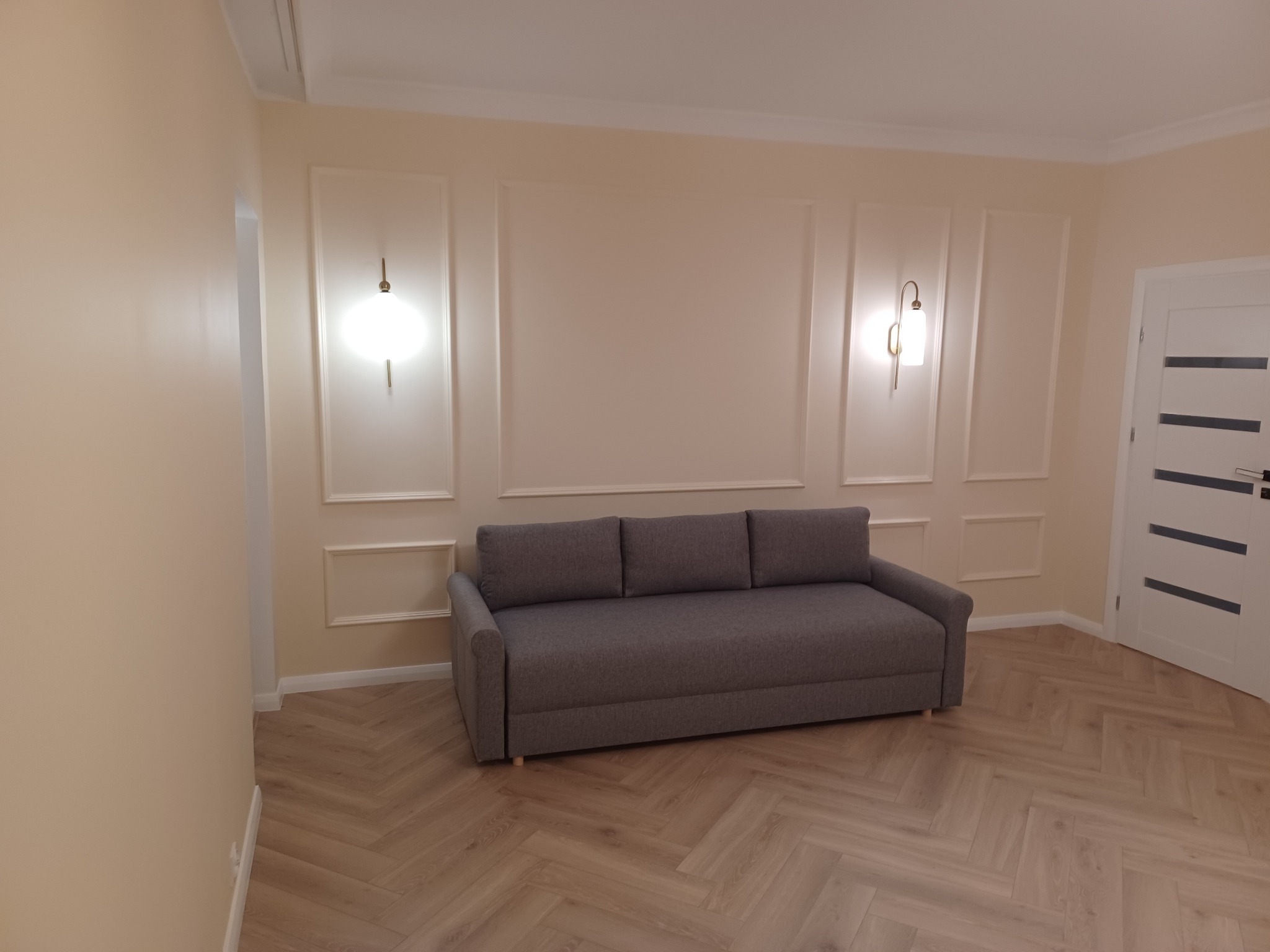 Jasny salon z szarą rozkładaną sofą, parkietem ułożonym w jodełkę, kremowymi ścianami z dekoracyjnymi listwami i kinkietami.