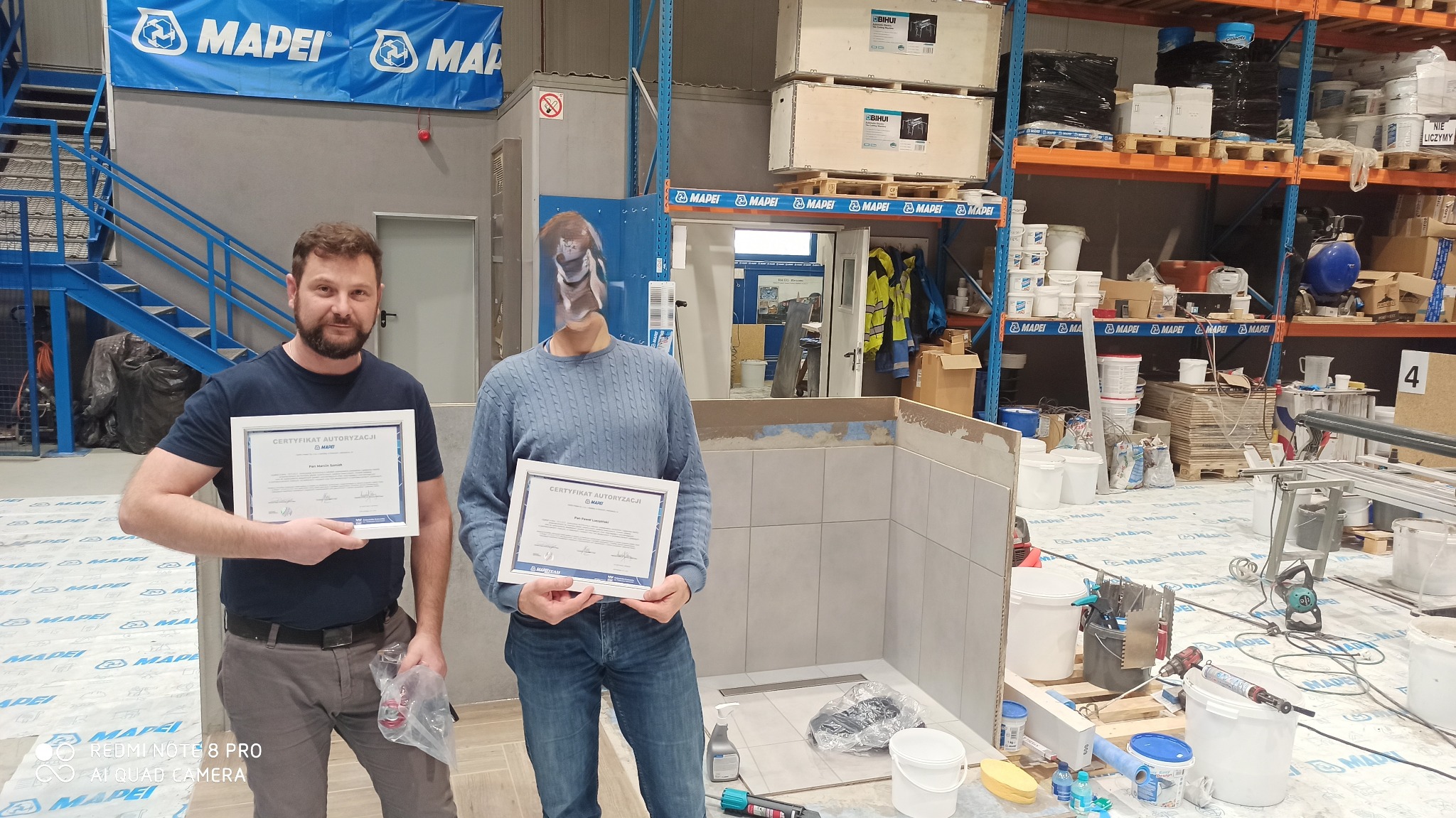 Dwóch mężczyzn prezentuje certyfikaty autoryzacji Mapei na tle częściowo wykończonej łazienki z szarymi płytkami, widoczny odpływ liniowy, narzędzia glazurnicze i materiały budowlane, podłoga...