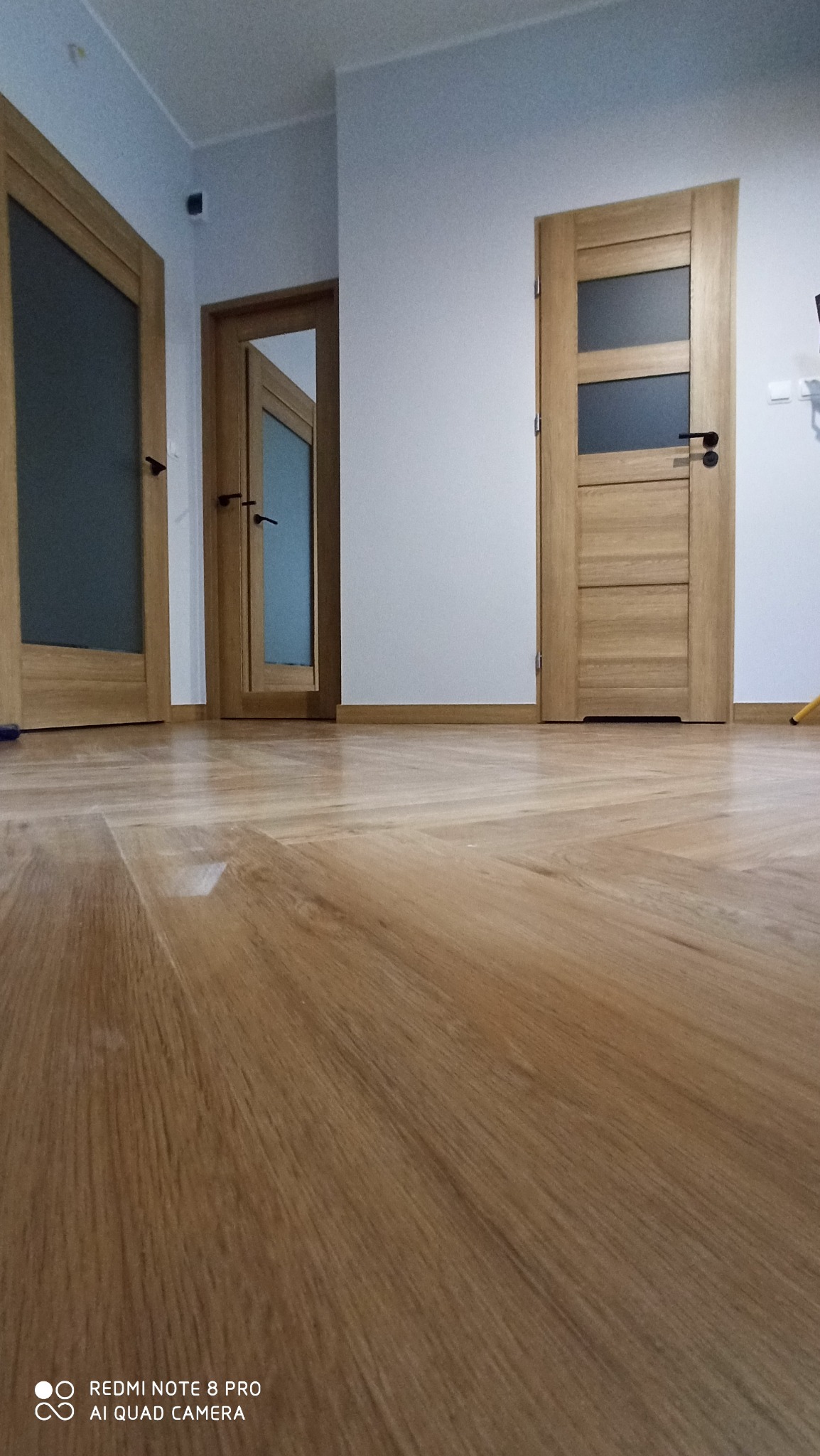 Ułożona parkietowa podłoga w jodełkę francuską, widoczne trzy drewniane drzwi z przeszkleniami i czarnymi klamkami, białe ściany i listwy przypodłogowe.