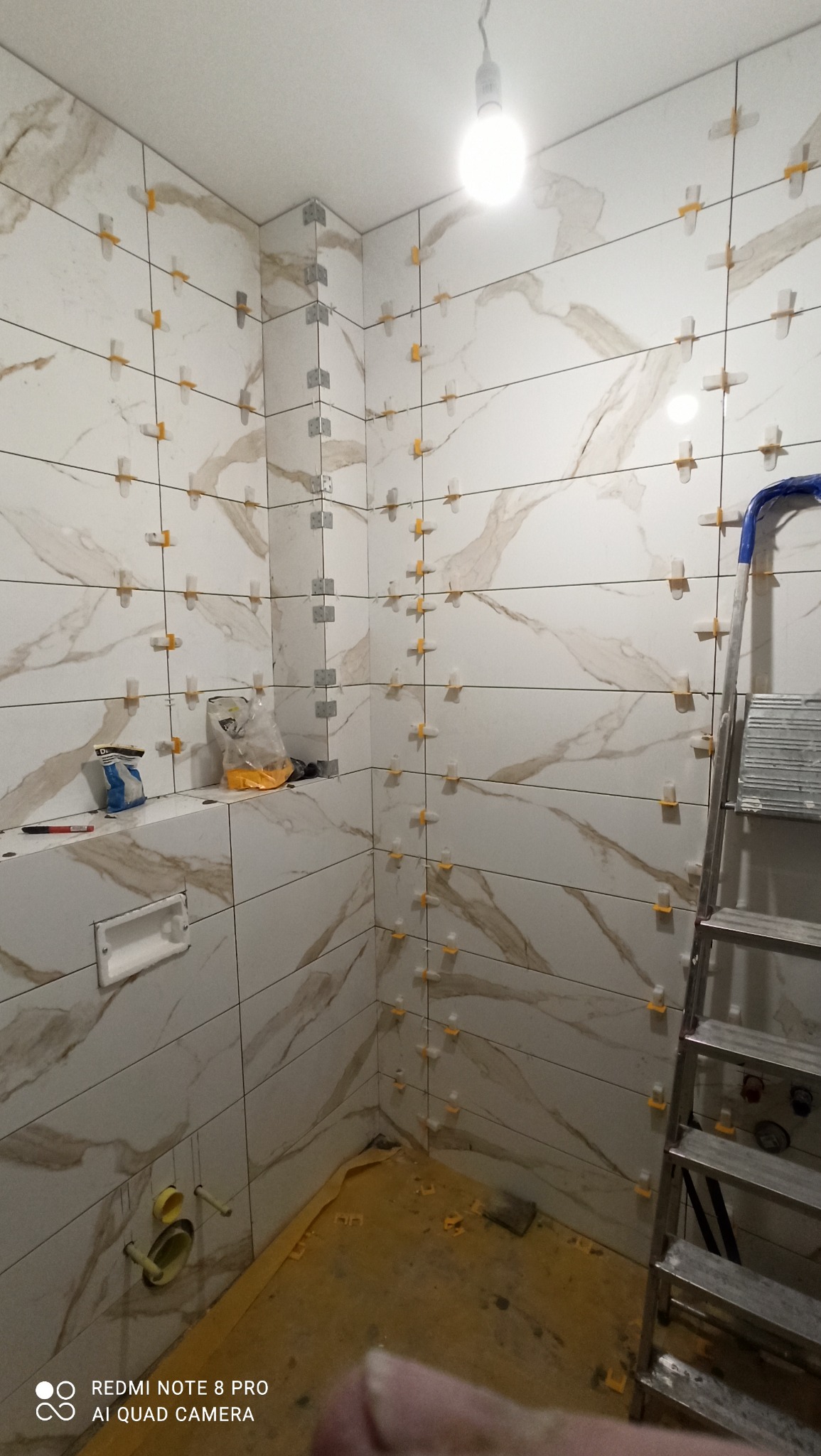 Łazienka w trakcie układania płytek marmuropodobnych z użyciem krzyżyków dystansowych i listew narożnych. Widoczna drabina i narzędzia glazurnicze.