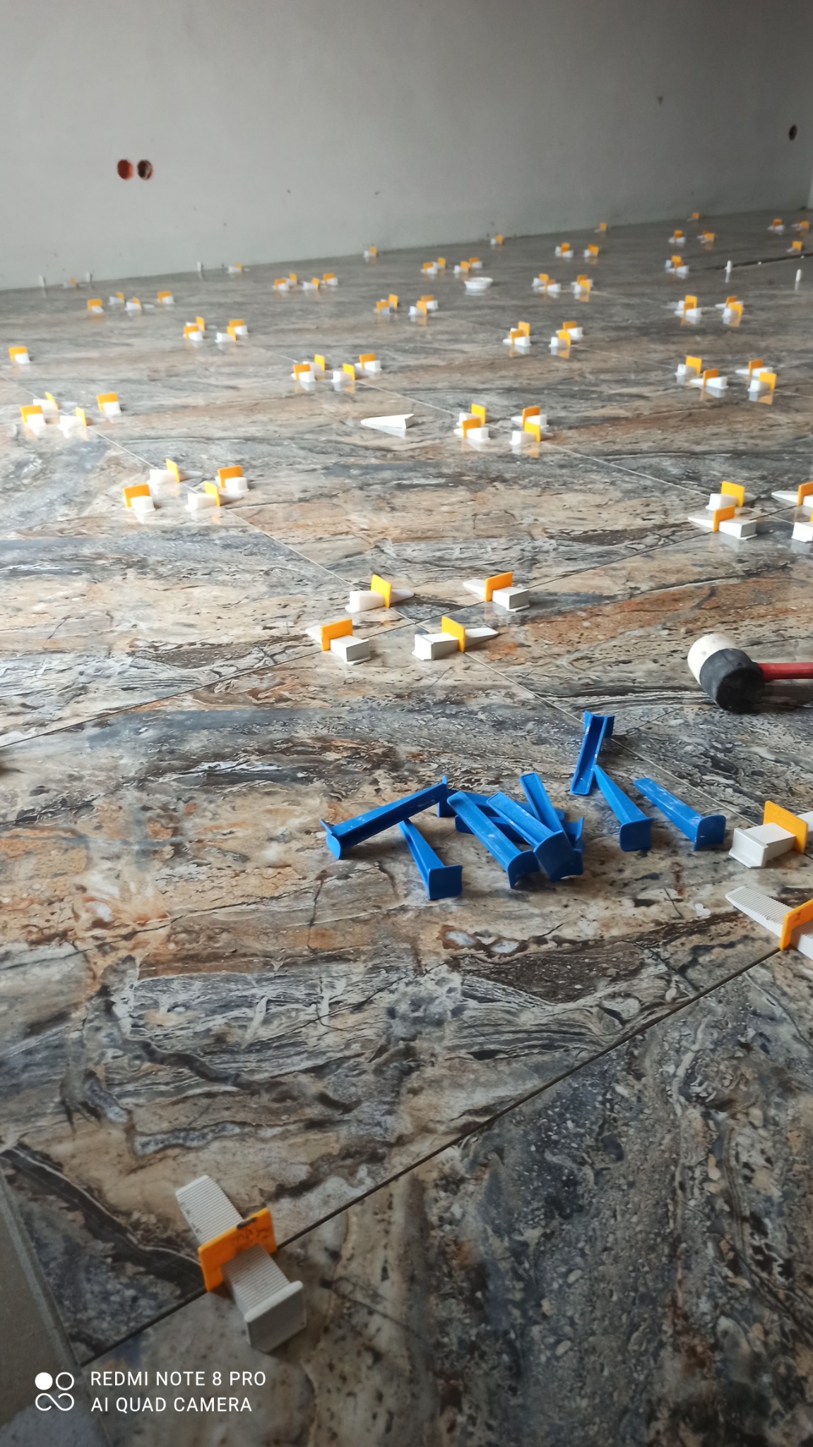 Układanie płytek podłogowych z użyciem systemu poziomowania. Widoczne kliny, klipsy i młotek gumowy. Płytki w odcieniach szarości i brązu z wyraźnym wzorem.