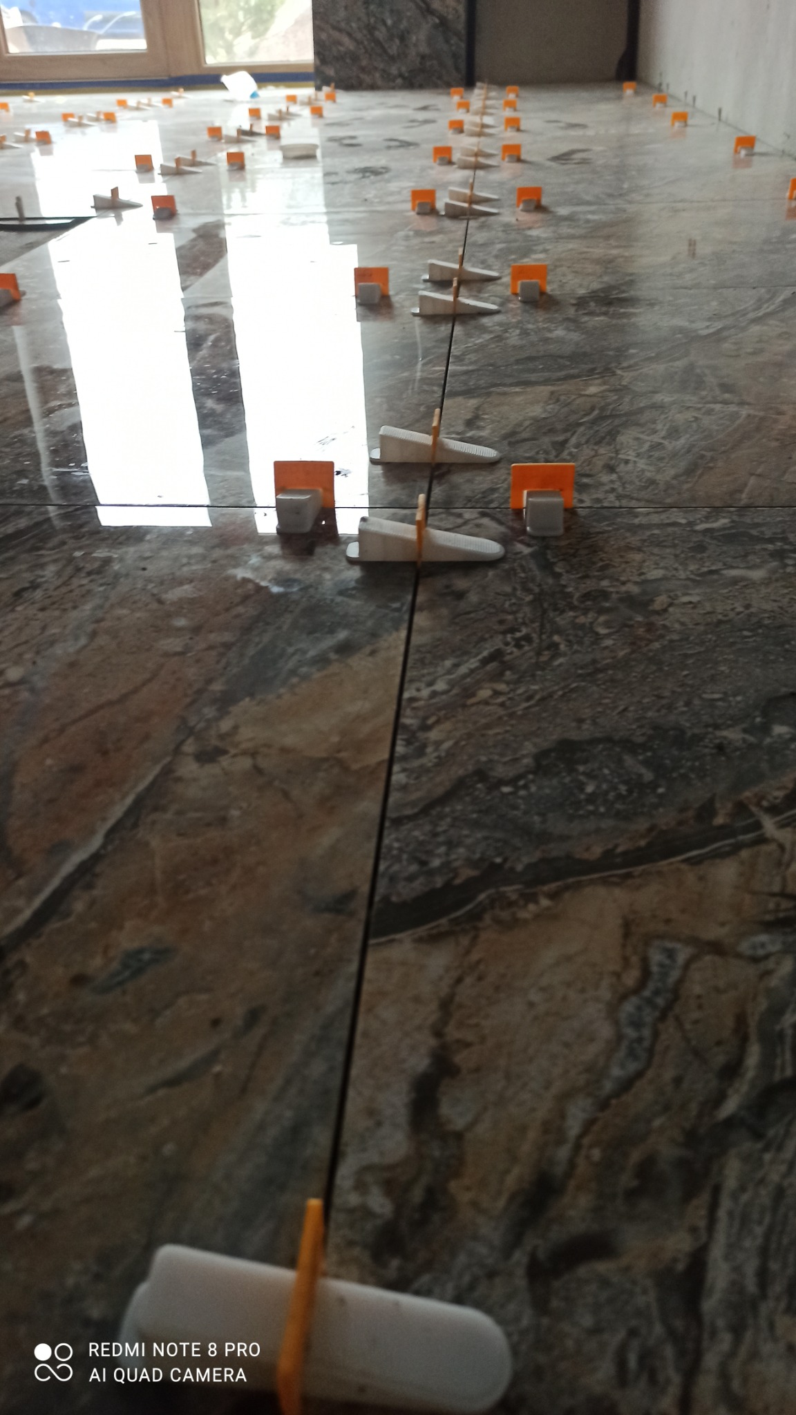 Połyskujące, marmurowe płytki podłogowe z systemem poziomowania płytek. Widoczne pomarańczowe kliny i białe klipsy. Płytki ułożone równo, z zachowaniem spoin.