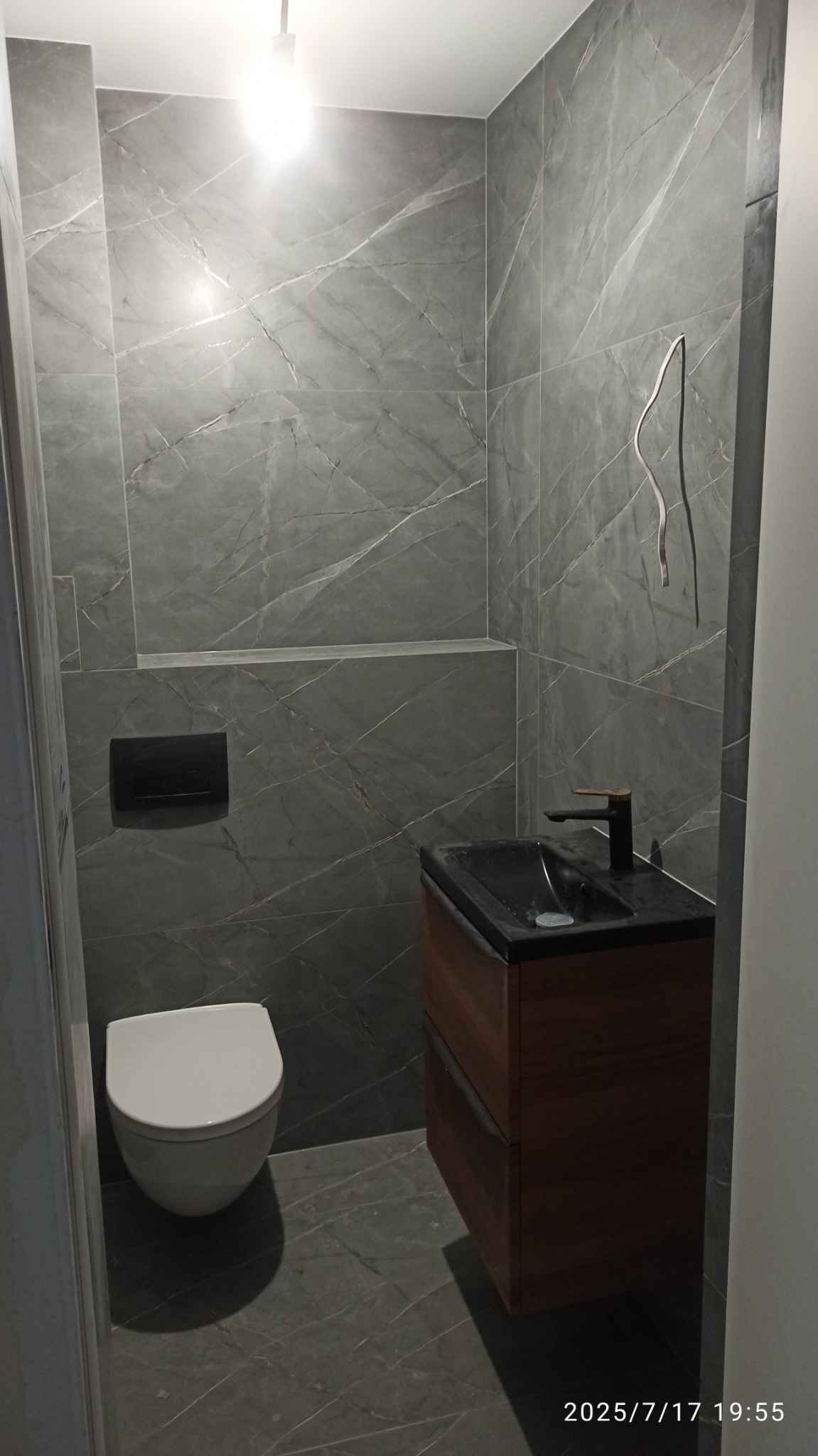 Nowoczesna łazienka z szarymi płytkami imitującymi marmur, podwieszana toaleta i drewniana szafka z czarną umywalką. W tle wnęka w ścianie.