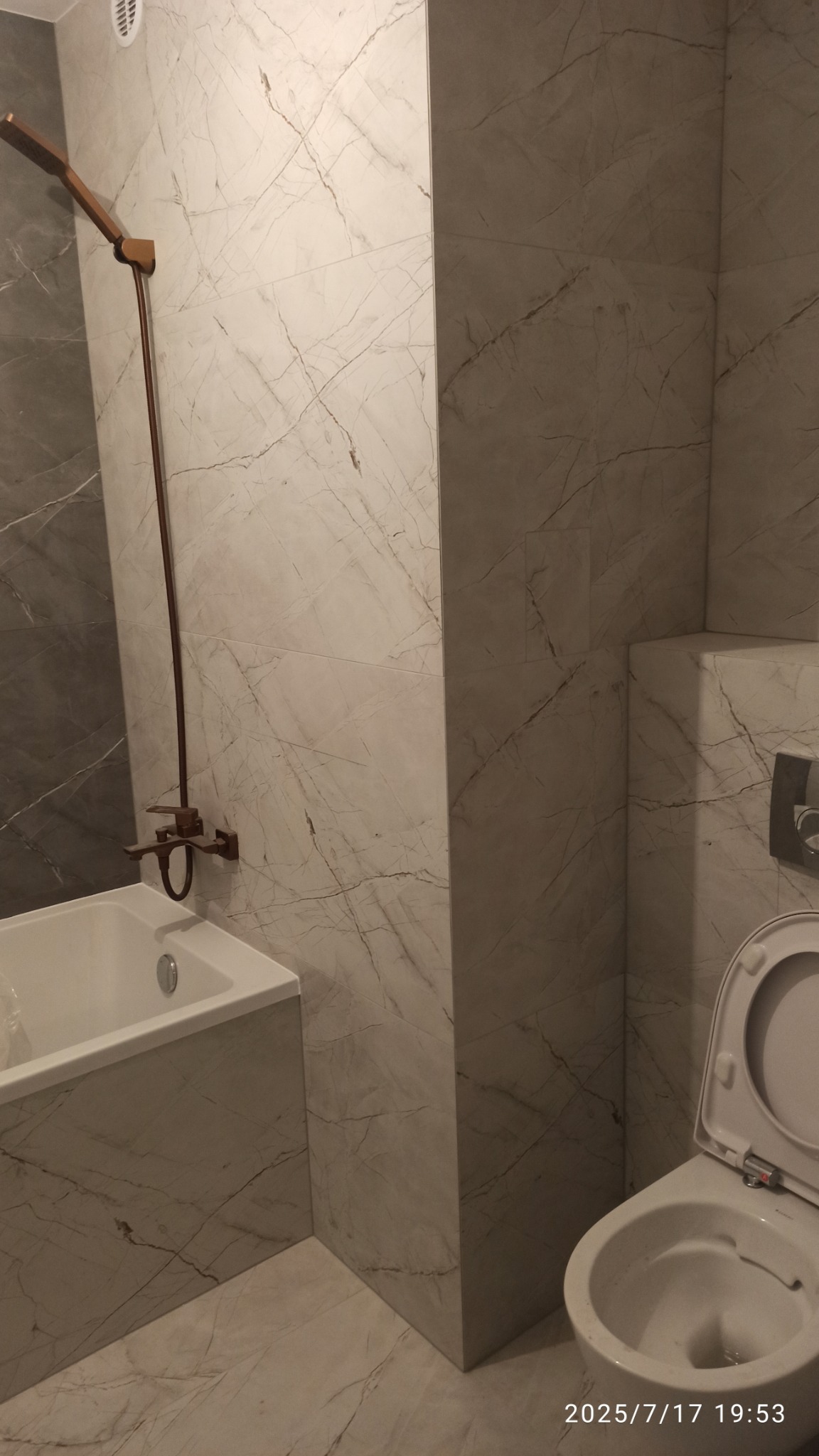 Wykończona łazienka z marmurowymi płytkami, brązową armaturą i białą ceramiką. Widoczna wanna, toaleta i bateria prysznicowa. Całość utrzymana w stonowanej kolorystyce.