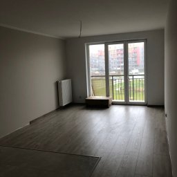 ProConcept - Jasne panele podłogowe w nowym mieszkaniu z grzejnikiem i balkonem z widokiem na blok. Świeżo wykończone ściany w odcieniach bieli i szarości.