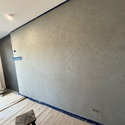 ProConcept - Ściana po szpachlowaniu dekoracyjnym w odcieniach szarości, zabezpieczona taśmą malarską. Na podłodze folia ochronna i stołek. Widoczne gniazdka elektryczne.