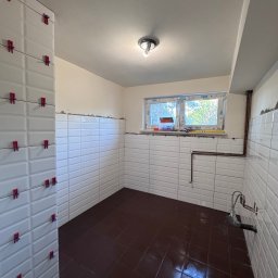 ProConcept - Łazienka w trakcie wykańczania: białe, prostokątne płytki na ścianach z czerwonymi klipsami poziomującymi, ciemnobrązowe płytki podłogowe, okno z widokiem na zieleń.