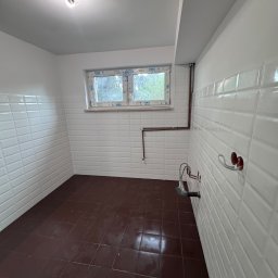 ProConcept - Wykończona łazienka z brązową podłogą i białymi, wypukłymi płytkami na ścianach. Widoczne rury i instalacja wodna. Okno z widokiem na zieleń.