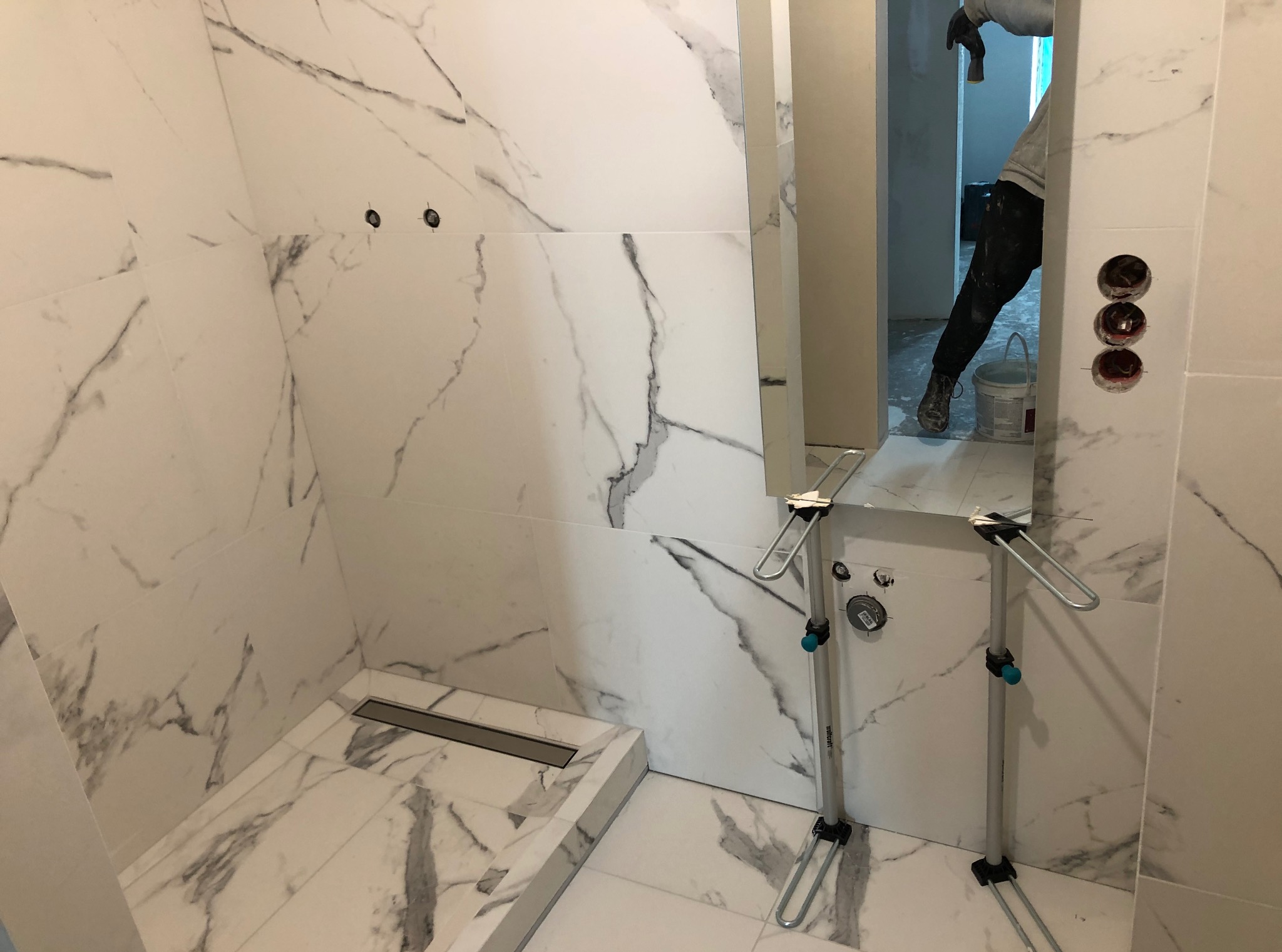 Wykończona łazienka w Bielawie z białymi płytkami imitującymi marmur, odpływem liniowym i narzędziami glazurniczymi w tle, częściowo widoczny pracownik.