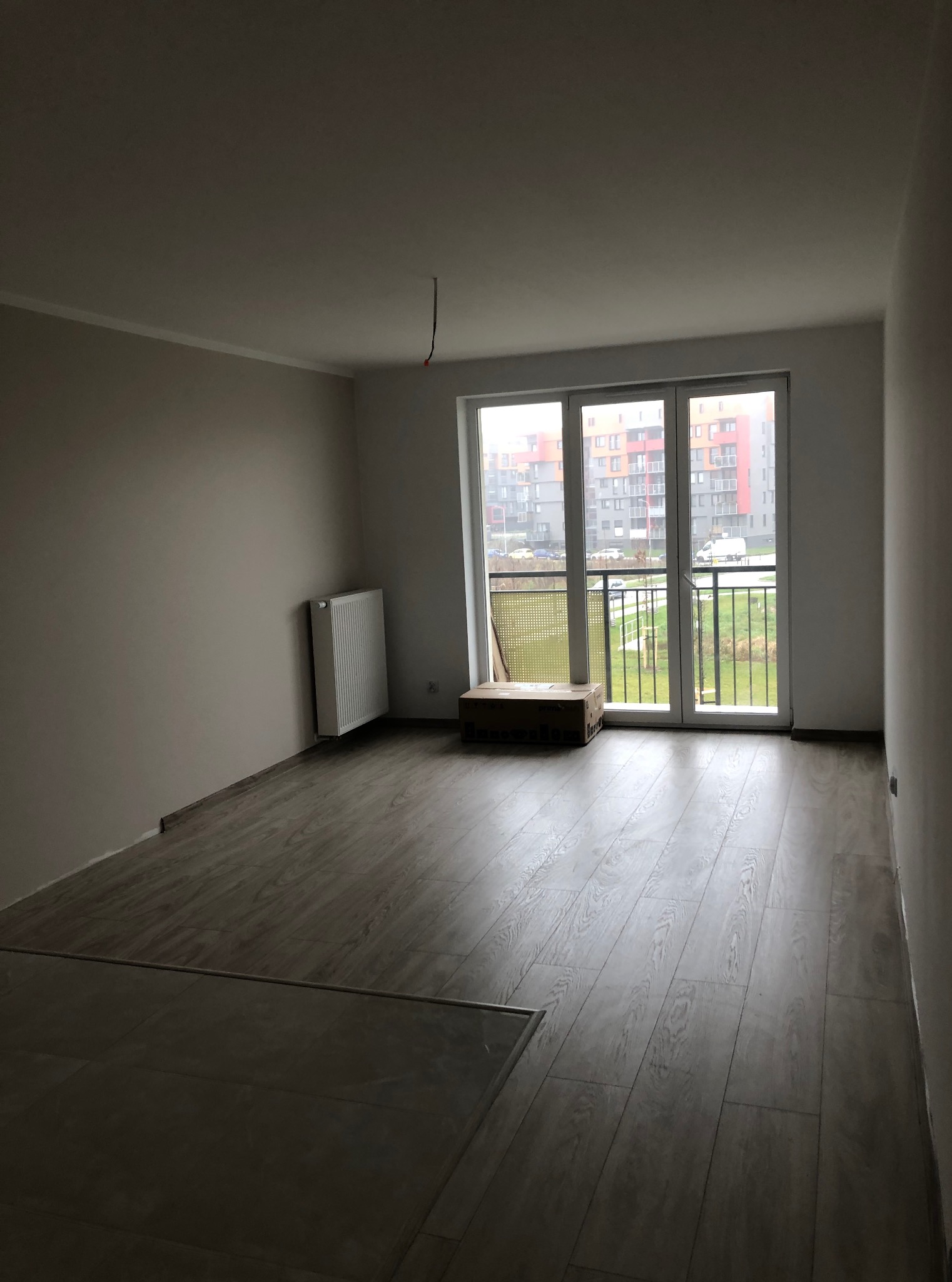 Jasne panele podłogowe w nowym mieszkaniu z grzejnikiem i balkonem z widokiem na blok. Świeżo wykończone ściany w odcieniach bieli i szarości.