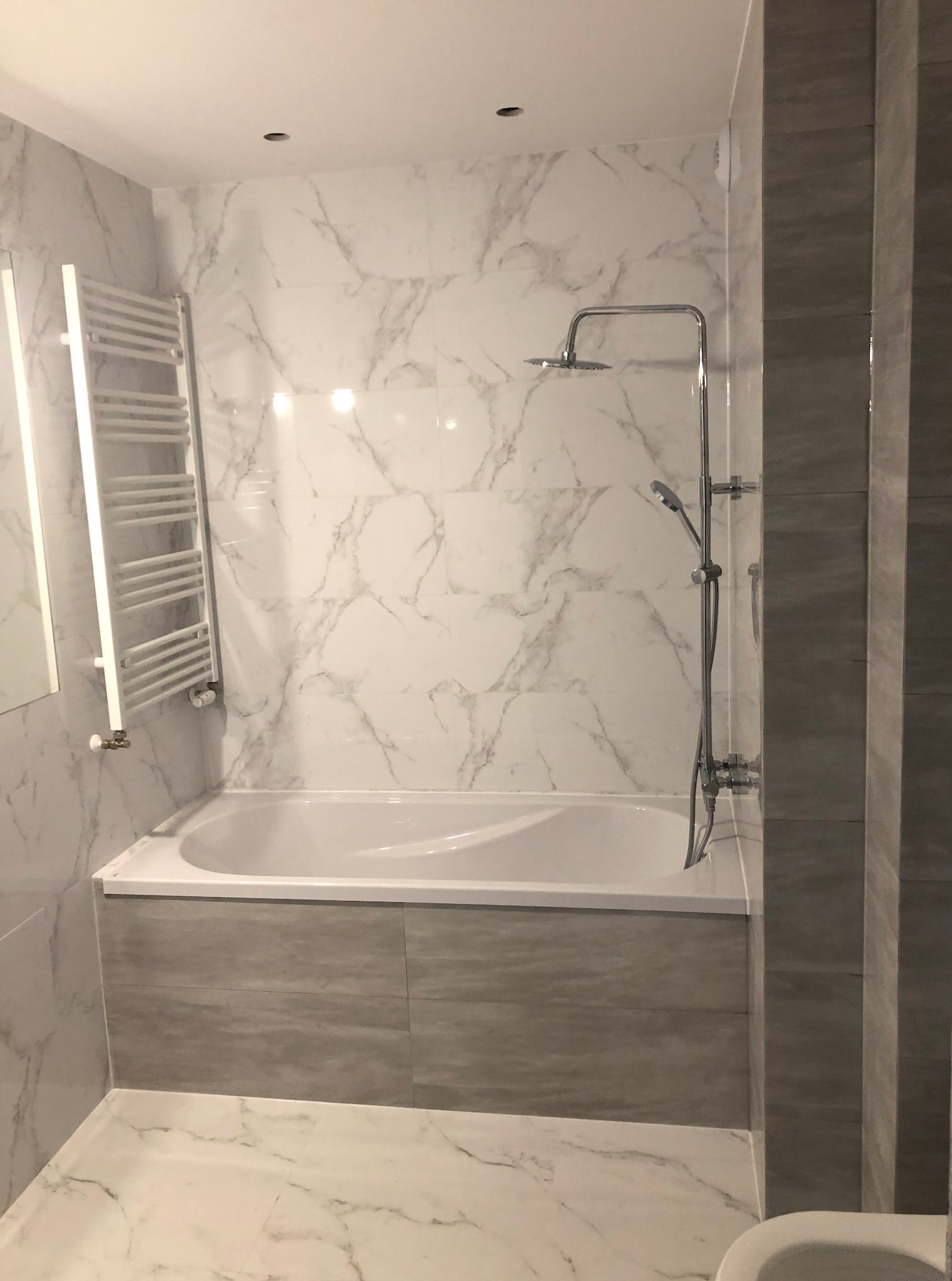 Wykończona łazienka z wanną obudowaną szarymi płytkami, białe marmurowe płytki na ścianach, grzejnik drabinkowy i chromowana armatura.