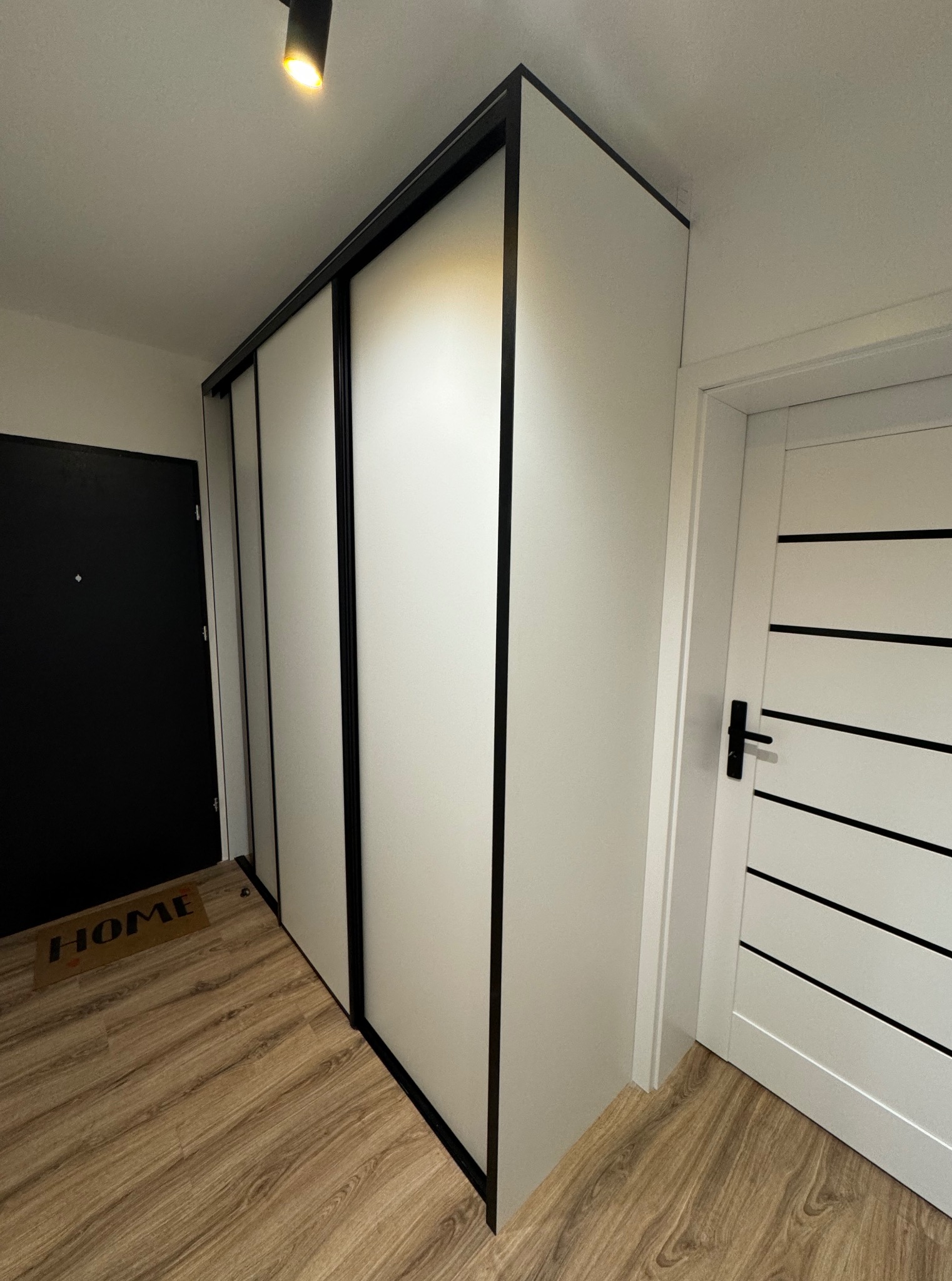 Nowoczesna szafa przesuwna z czarnymi obramowaniami w jasnym przedpokoju, widoczna z perspektywy podłogi, obok drzwi z poziomymi, czarnymi paskami.