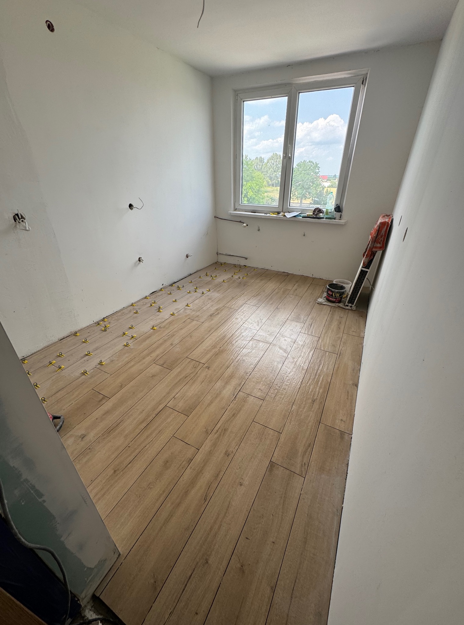 Układanie drewnopodobnych płytek podłogowych w jasnym pokoju z oknem. Żółte krzyżyki dystansowe między płytkami. Widoczne narzędzia i materiały budowlane.