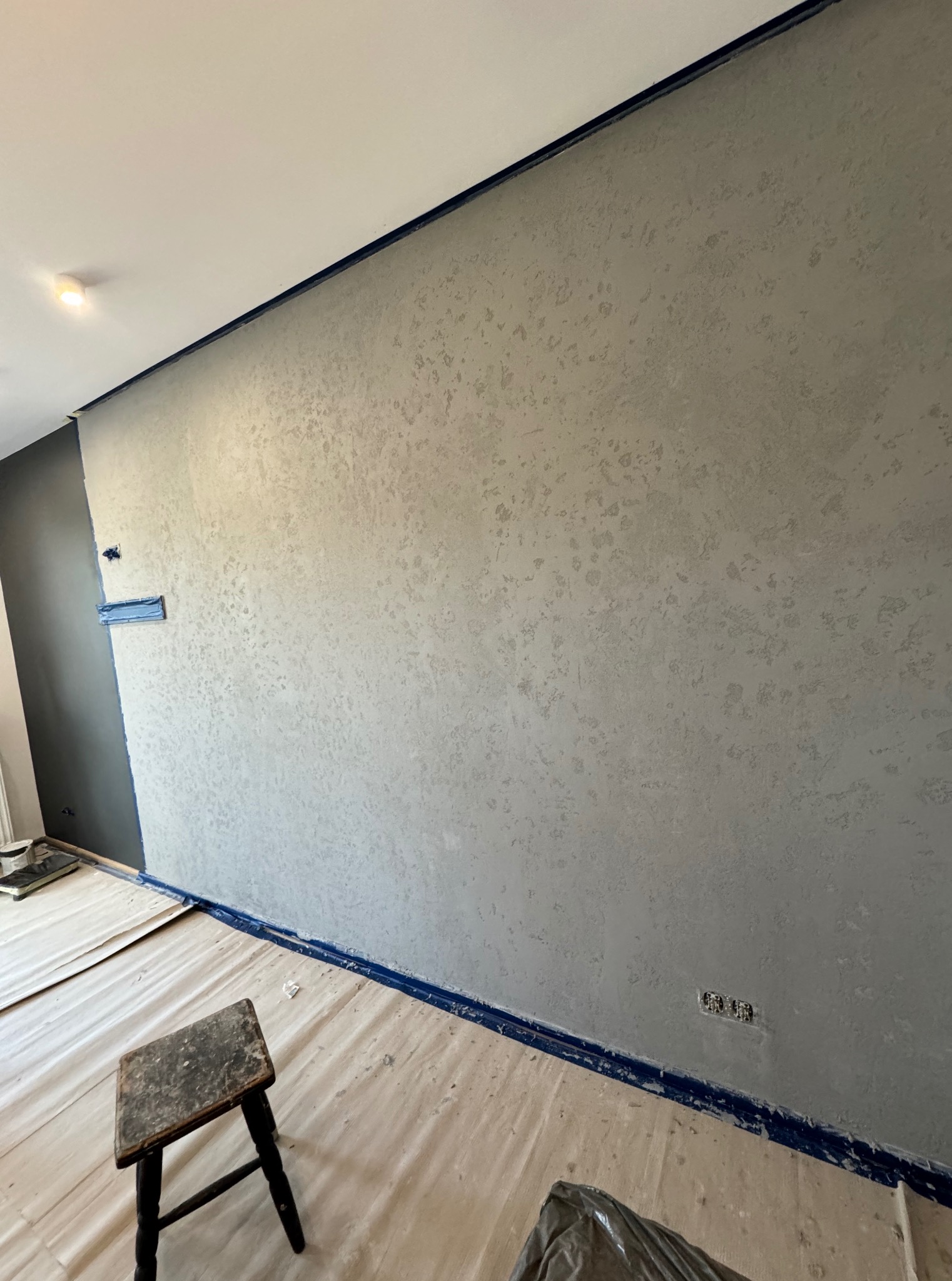 Ściana po szpachlowaniu dekoracyjnym w odcieniach szarości, zabezpieczona taśmą malarską. Na podłodze folia ochronna i stołek. Widoczne gniazdka elektryczne.