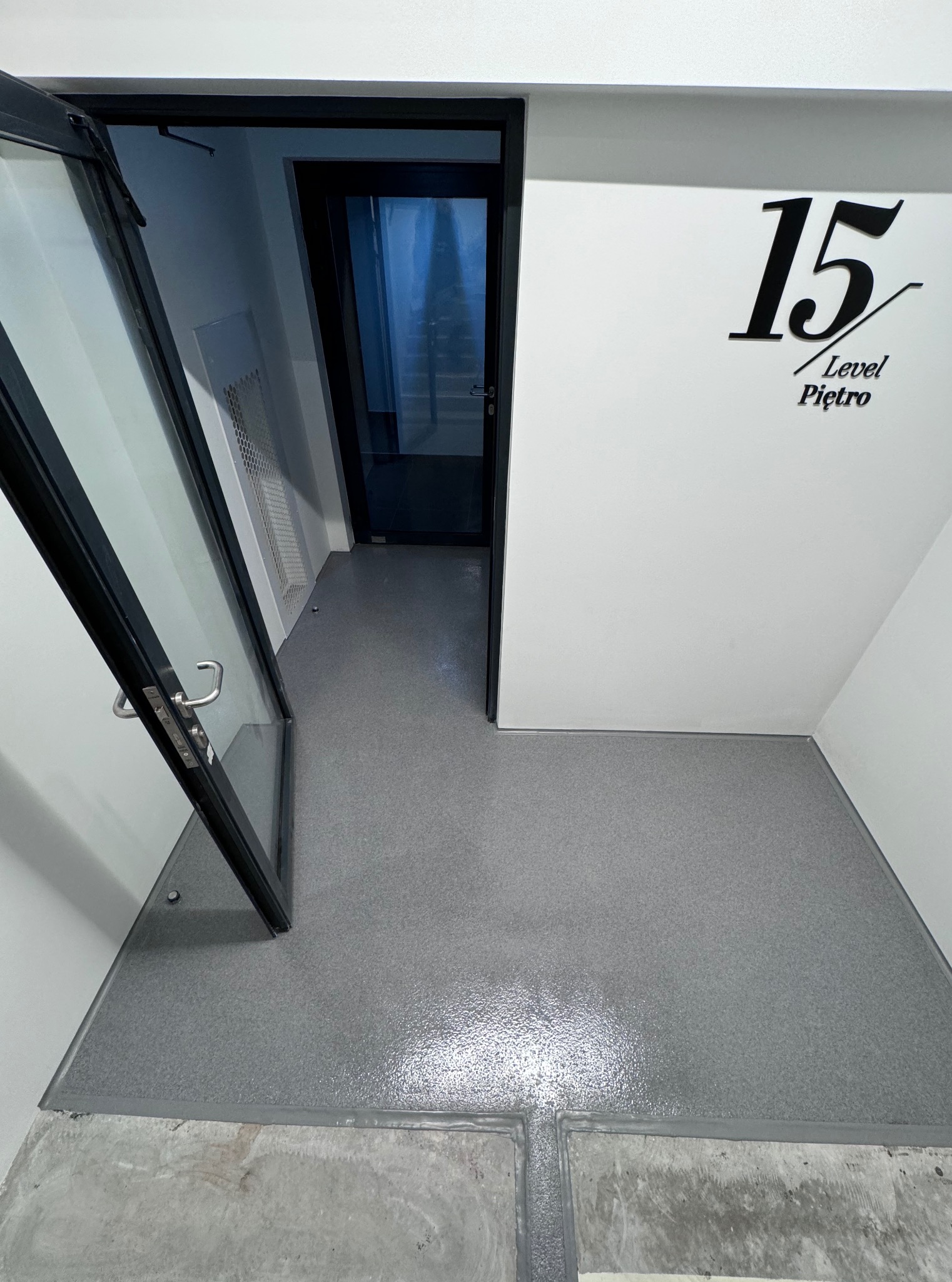 Szare, błyszczące wykończenie podłogi w korytarzu z otwartymi, przeszklonymi drzwiami i numerem 15 na ścianie. Widok z góry, nowoczesny design.