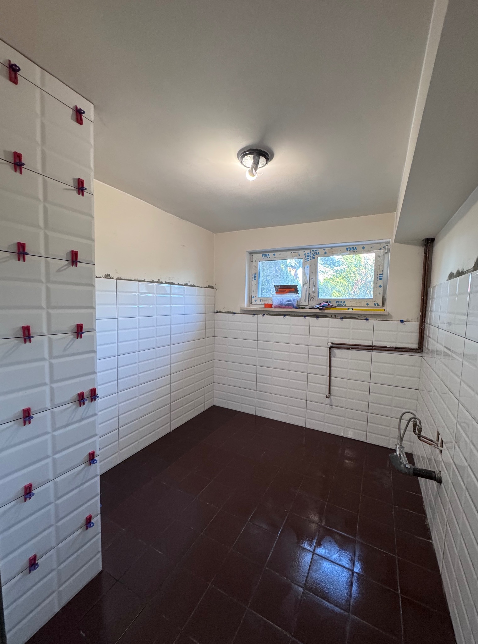 Łazienka w trakcie wykańczania: białe, prostokątne płytki na ścianach z czerwonymi klipsami poziomującymi, ciemnobrązowe płytki podłogowe, okno z widokiem na zieleń.