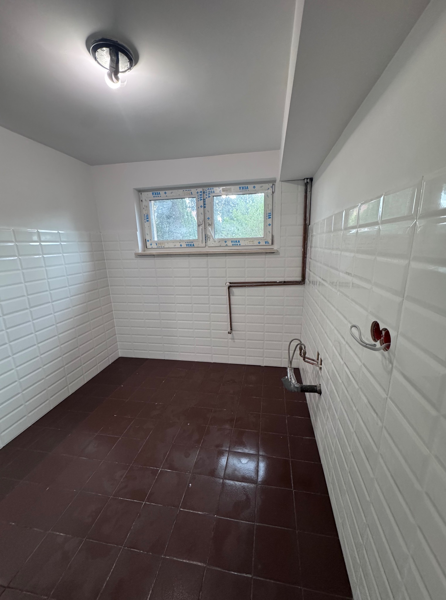 Wykończona łazienka z brązową podłogą i białymi, wypukłymi płytkami na ścianach. Widoczne rury i instalacja wodna. Okno z widokiem na zieleń.