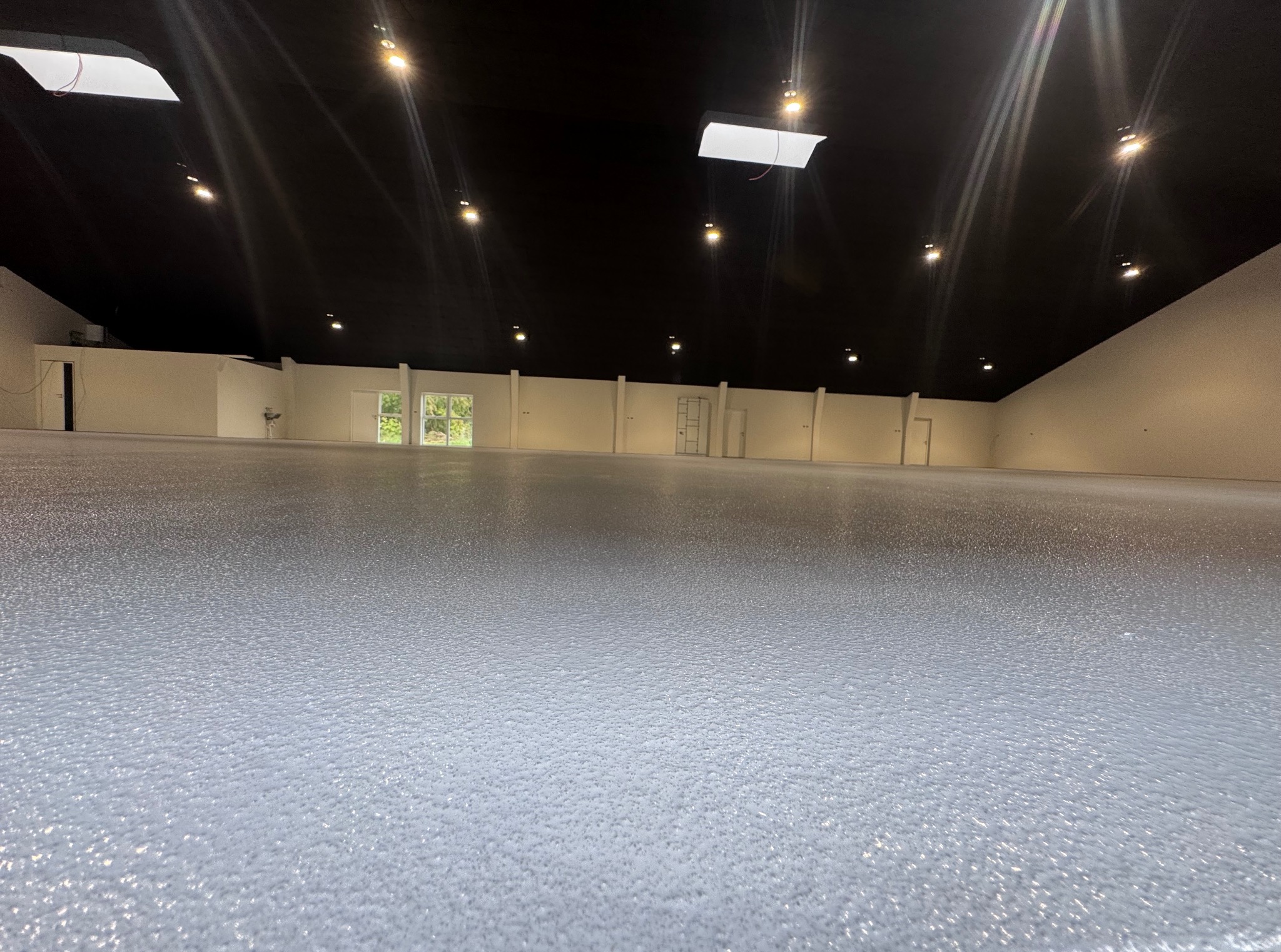 Błyszcząca, szara posadzka w przestronnym, jasnym pomieszczeniu z czarnym sufitem i rzędem okien w tle. Widoczne punkty świetlne na suficie.