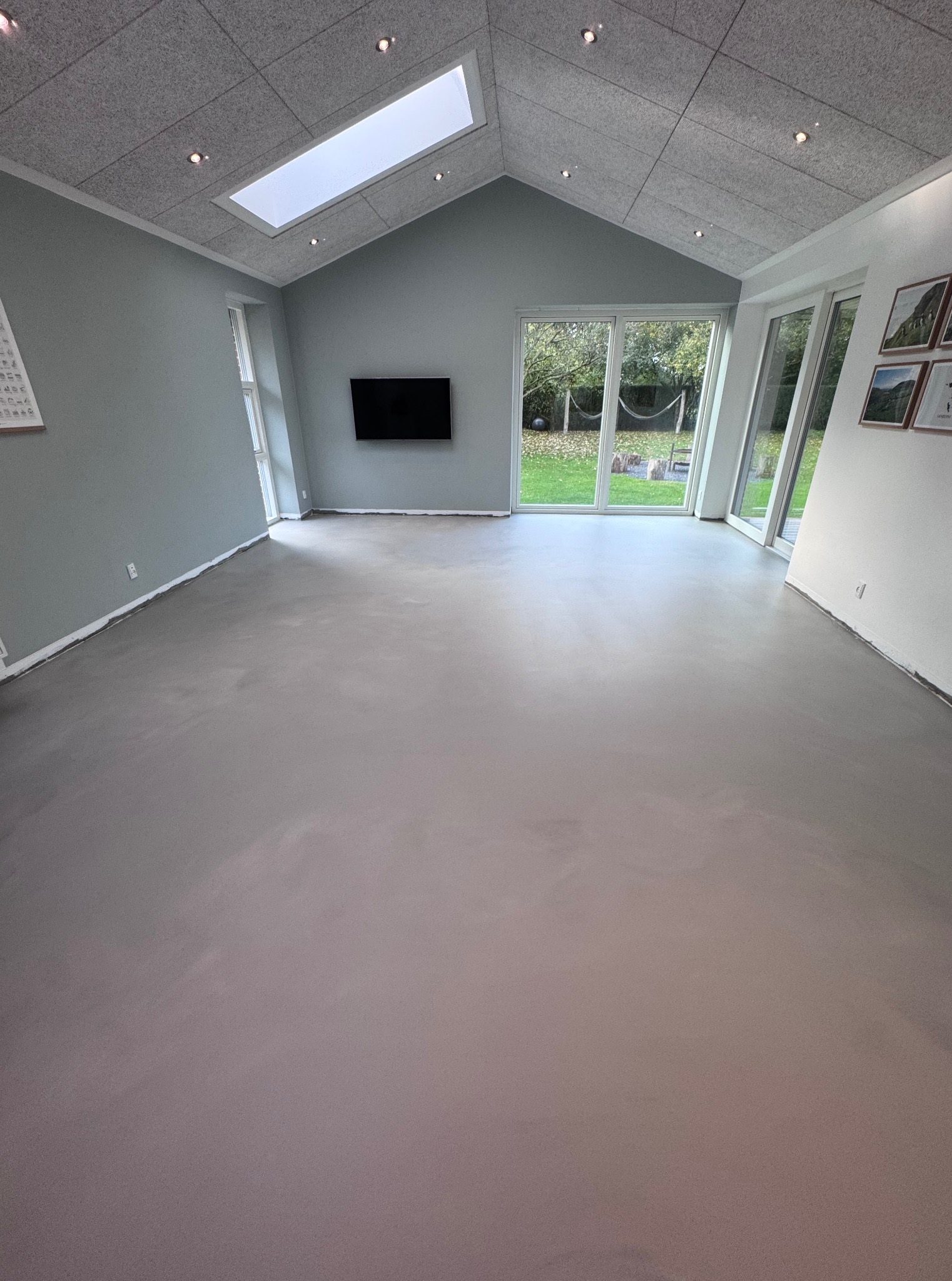 Jasnoszara wylewka samopoziomująca w przestronnym pokoju z oknami na ogród i sufitem z panelami akustycznymi oraz oknem dachowym. Minimalistyczny design.