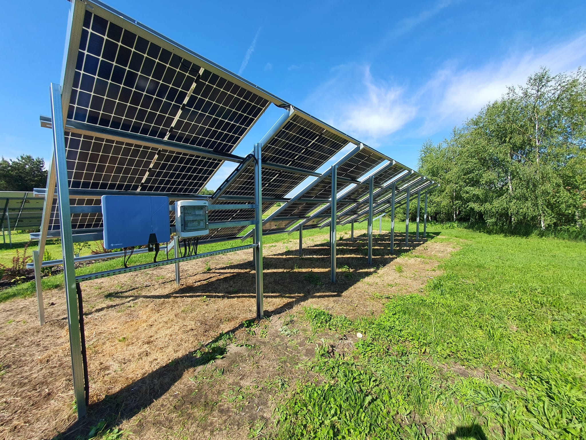 Rząd paneli słonecznych zamontowanych na metalowej konstrukcji na trawiastym terenie, z widocznymi inwerterami i błękitnym niebem w tle.