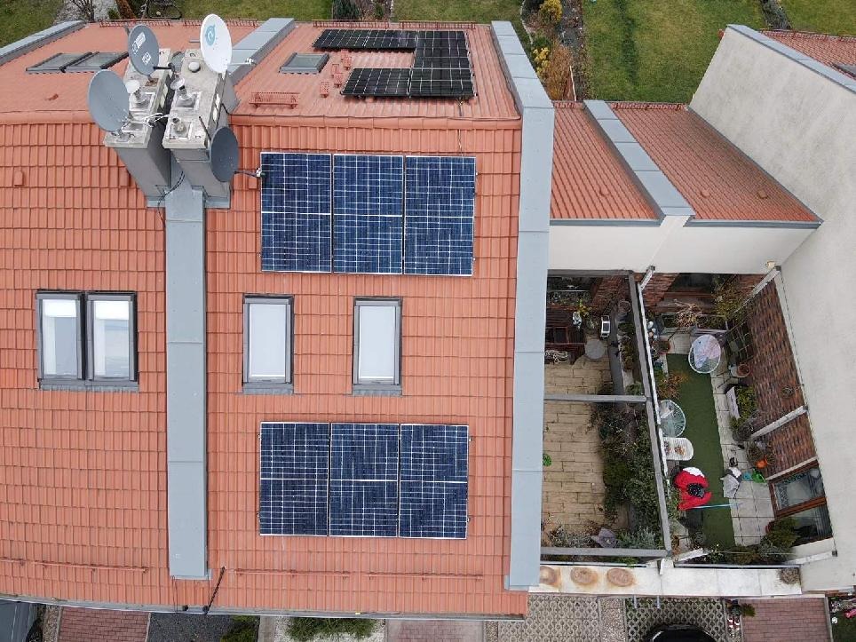 Widok z góry na dom z czerwoną dachówką, na której zamontowano panele fotowoltaiczne oraz anteny satelitarne, widoczny również mały ogródek.