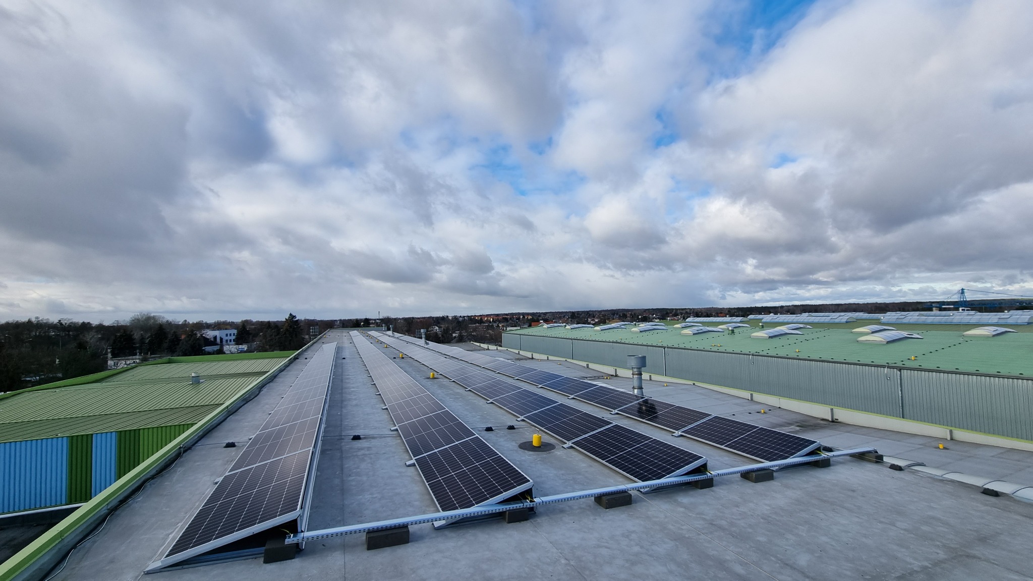 Rzędy paneli słonecznych na dachu przemysłowym z widokiem na miasto pod pochmurnym niebem.