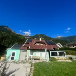 Dom jednorodzinny z panelami słonecznymi na dachu z czerwonej dach&oacute;wki, drabina oparta o ścianę, w tle g&oacute;ry i błękitne niebo.