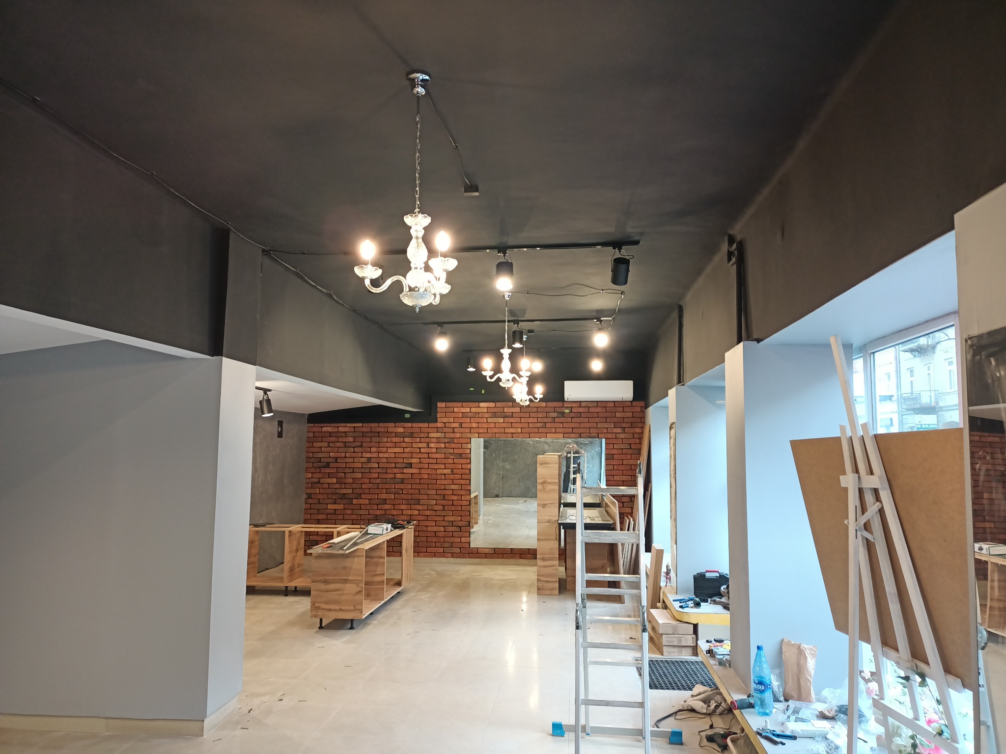Wnętrze lokalu w trakcie prac wykończeniowych: widoczne podwieszane lampy, ściana z cegły, drabina, drewniane konstrukcje meblowe i sztaluga z płytą.